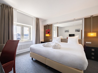 Moderne hotelkamer met een groot bed, wit beddengoed, nachtlampen en een raam met stadsuitzicht. De kamer biedt een comfortabele en uitnodigende sfeer, perfect voor ontspanning.