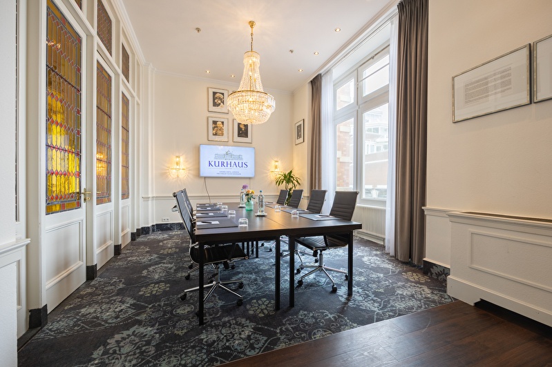 Vergaderruimte Paul van Vliet met een tafel, leren stoelen, kristallen kroonluchter en een scherm met het Kurhaus-logo. Grote ramen en glas-in-looddeuren zorgen voor veel lichtinval.