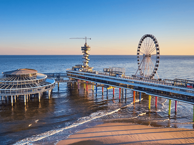 The pier in Scheveningen with a Ferris wheel and a beach in the background, under a blue sky.