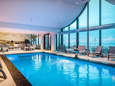 Wellness area with an indoor pool, sun loungers and large windows overlooking the sea. Furnishings include plants and a wall decoration with a historical scene.