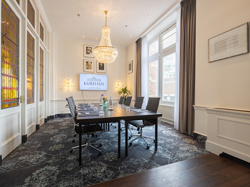 Meeting room with a table, leather chairs, crystal chandelier, and a screen with the Kurhaus logo. Large windows and stained-glass doors provide plenty of natural light.