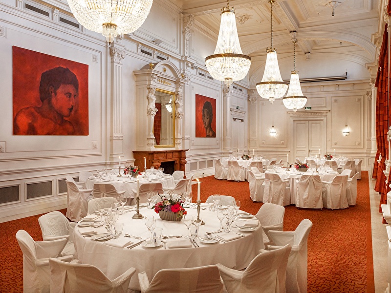 Elegant meeting room with round, white-covered tables, crystal chandeliers, and classic white walls with red artworks.