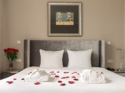 Elegant hotelbed met witte lakens, met rode rozenblaadjes en badjassen. Naast het bed staat een vaas met rode rozen, en aan de muur hangt een kunstafbeelding.